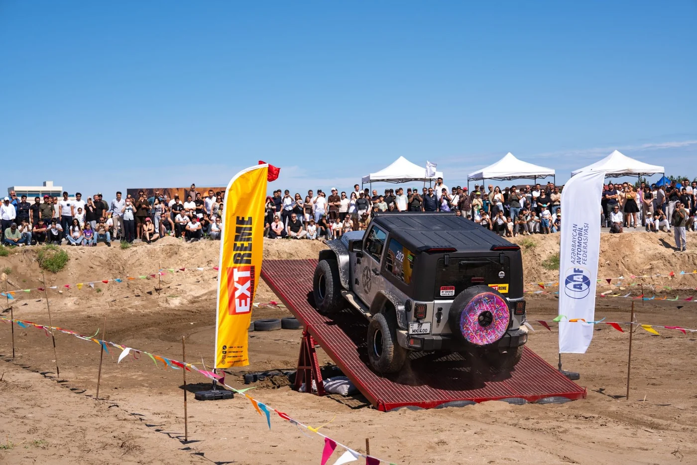 An off-road vehicle drives onto the ramp platform, with spectators and tents behind the markings